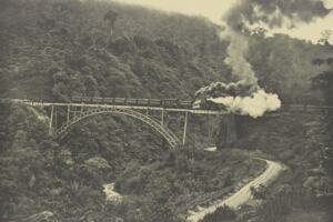 A sepia-toned photograph depicting a historic steam locomotive billows dramatic clouds of smoke as it traverses a steel bridge, suspended above a lush, forested valley of Dutch East Indies.